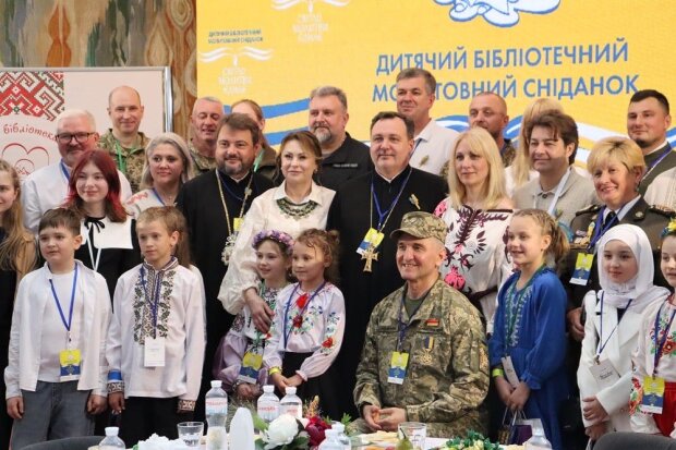 На фото група людей, серед яких діти та дорослі, зібралися на заході. Позаду них жовтий банер із синім текстом: «Дитячий бібліотечний мотиваційний сніданок». Дехто у військовій формі, інші – у традиційному одязі. На столі перед ними стоять пляшки з водою, квіти та інші предмети.