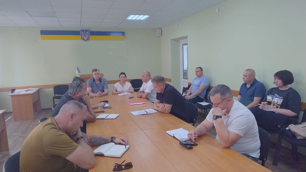  Група людей сидить за великим столом у кімнаті з прапором України та гербом на стіні. Учасники беруть участь у нараді — на столі лежать документи, блокноти, ручки та смартфони. Атмосфера офіційна, інтер'єр — світло-зелений із білою стелею та освітленням.