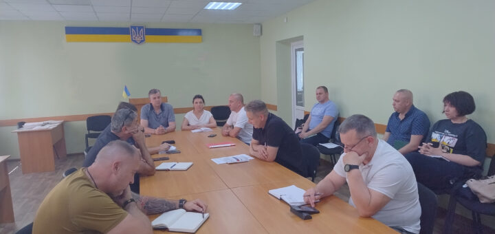 Група людей сидить за великим столом у кімнаті з прапором України та гербом на стіні. Учасники беруть участь у нараді — на столі лежать документи, блокноти, ручки та смартфони. Атмосфера офіційна, інтер'єр — світло-зелений із білою стелею та освітленням.