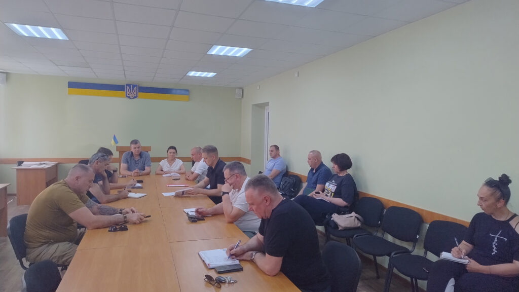  Офіційна зустріч у приміщенні з українським прапором і гербом на стіні. За великим прямокутним столом сидить кілька людей — вони спілкуються, роблять нотатки й обговорюють документи. Освітлення яскраве, у кімнаті — світло-зелені стіни й додаткові стільці вздовж них.