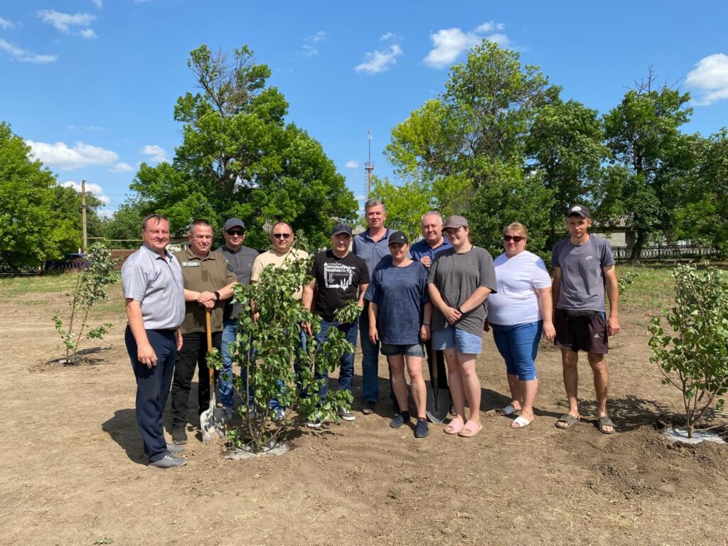 Ця фотографія передає дух єдності та турботи про природу. На ній зображена група людей, які стоять на відкритому полі серед молодих дерев. Дехто тримає лопати, що свідчить про їхню участь у висадці дерев. Навколо—зелена рослинність, дерев’яний паркан і яскраве блакитне небо з кількома хмарами.