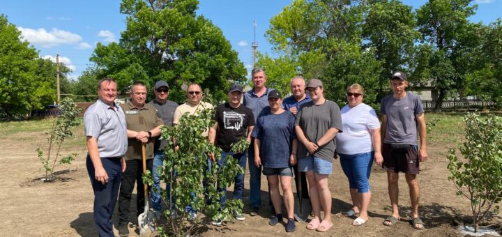 Ця фотографія передає дух єдності та турботи про природу. На ній зображена група людей, які стоять на відкритому полі серед молодих дерев. Дехто тримає лопати, що свідчить про їхню участь у висадці дерев. Навколо—зелена рослинність, дерев’яний паркан і яскраве блакитне небо з кількома хмарами.