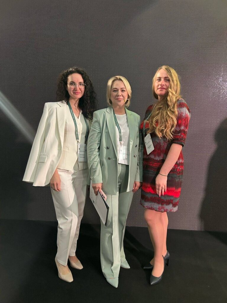 Троє людей стоять поруч на тлі темної текстурованої стіни. Зліва — жінка у білому костюмі з білою блузою та бежевими туфлями. У центрі — жінка у світло-зеленому костюмі з білою блузою, яка тримає папку. Справа — жінка у червоно-сірій сукні з чорними туфлями на підборах. Усі троє мають бейджі на ланцюжках.