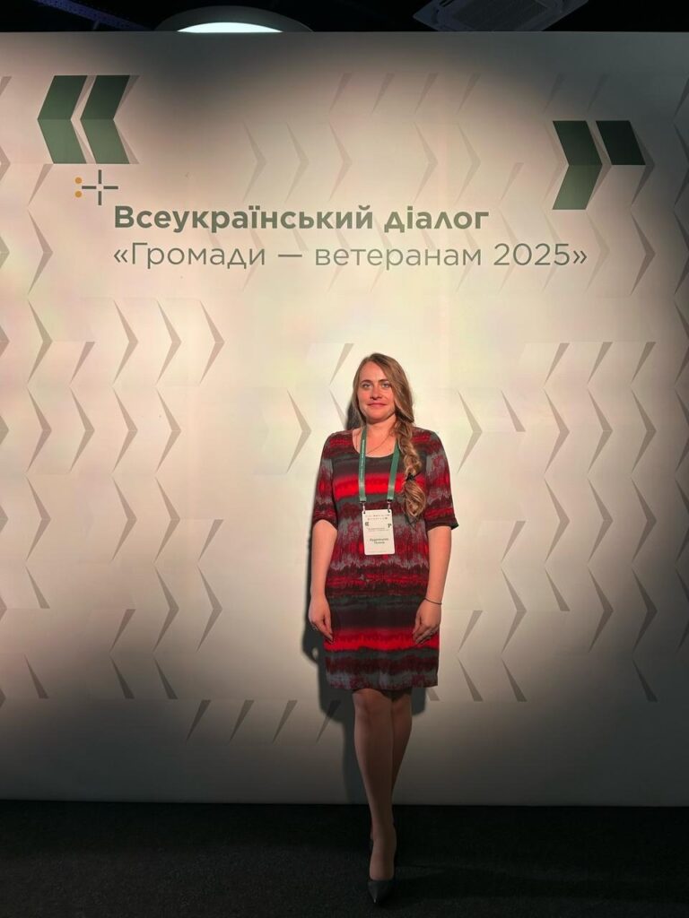 Галина Кудрявцева стоїть перед фоном із текстом українською мовою. Фон має геометричний малюнок зі стрілками, що вказують праворуч. Текст на фоні: "Всеукраїнський діалог «Громади — ветеранам 2025»