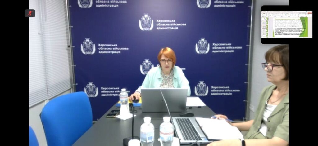 У конференц-залі Херсонської обласної військової адміністрації троє учасників сидять за столом із ноутбуками та документами. На екрані — слайд презентації з вимогами до проведення тренінгів з цивільного захисту. Атмосфера ділова, тло — синє з гербом і написом адміністрації.