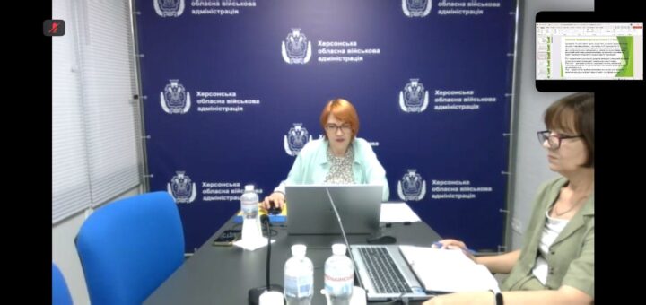 У конференц-залі Херсонської обласної військової адміністрації троє учасників сидять за столом із ноутбуками та документами. На екрані — слайд презентації з вимогами до проведення тренінгів з цивільного захисту. Атмосфера ділова, тло — синє з гербом і написом адміністрації.