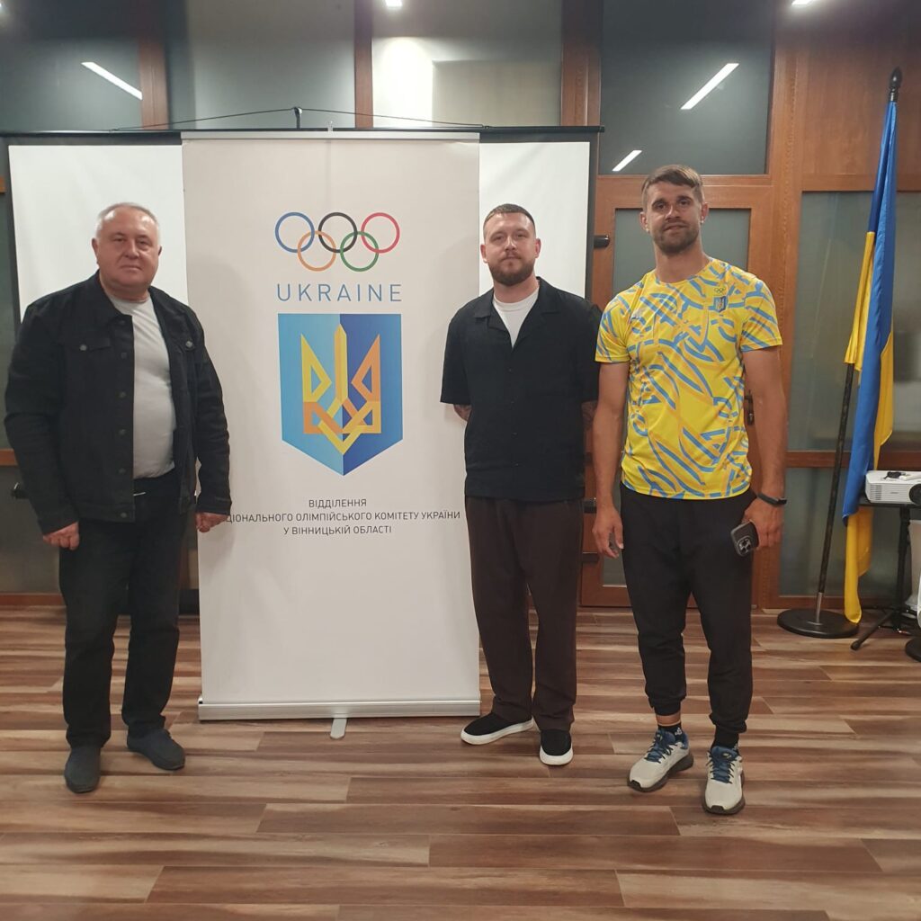 Три чоловіка стоять перед банером із логотипом Олімпійських кілець, написом «UKRAINE» і тризубом. Сергій Петроченко — ліворуч. Тло — синьо-жовтий прапор України. На банері вказано: «Відділення Національного олімпійського комітету України у Вінницькій області».