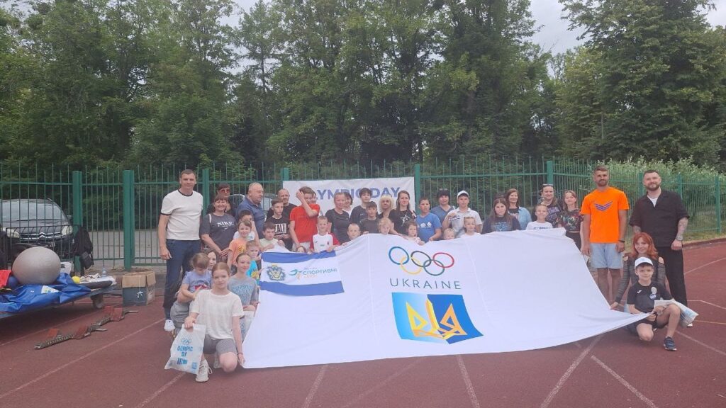 Групове фото дорослих і дітей на стадіоні під час заходу «Олімпійський день». Учасники тримають два банери: із написом «Olympic Day» та з логотипом Олімпійських кілець, тризубом і словом «Ukraine». Атмосфера святкова, позаду — дерева й огорожа.