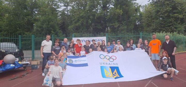 Групове фото дорослих і дітей на стадіоні під час заходу «Олімпійський день». Учасники тримають два банери: із написом «Olympic Day» та з логотипом Олімпійських кілець, тризубом і словом «Ukraine». Атмосфера святкова, позаду — дерева й огорожа.