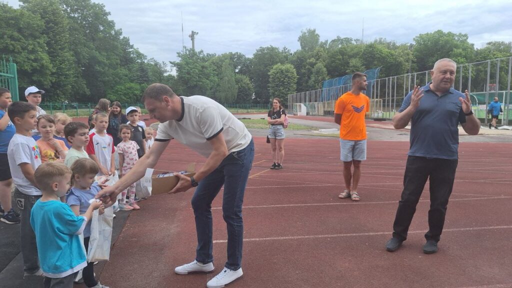 Діти стоять у шерензі на біговій доріжці стадіону, дорослий роздає їм предмети — ймовірно, подарунки або нагороди. Позаду — ще троє дорослих, один із них у помаранчевій футболці. Атмосфера — дружня, тло — зелені дерева та спортивний майданчик.