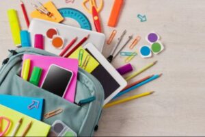 Рюкзак блакитного кольору зі шкільним приладдям: смартфон, планшет, блокнот, олівці, ручки, маркери, ножиці, циркуль, лінійка, клейкі нотатки й канцелярські дрібниці. Все лежить розсипом — сцена наче зібрана до навчання.