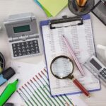 На столі — канцелярське приладдя, калькулятори, графіки, блокнот і чашка кави. Видно аркуш із числами, лупу та олівець. Сцена нагадує робоче місце для фінансового або аналітичного обліку