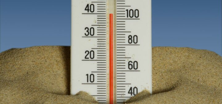 На зображенні — термометр, частково занурений у пісок. Він має дві шкали: ліворуч — за Цельсієм (від -10 до 50 градусів), праворуч — за Фаренгейтом (від 20 до 120 градусів). Рівень червоної рідини показує приблизно 30°C або 86°F. На тлі — ясне синє небо, що створює враження спекотного дня на пляжі або в пустелі.