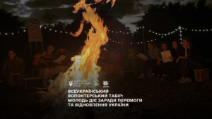 Група людей сидить навколо великого багаття вночі. Вогонь яскраво горить, освітлюючи простір. Деякі учасники тримають папери, один грає на гітарі. На фоні — дерева та гірлянди зі світлами, що створюють затишну атмосферу. Угорі — текст українською: «МІНІСТЕРСТВО МОЛОДІ ТА СПОРТУ УКРАЇНИ. ВСЕУКРАЇНСЬКИЙ ВОЛОНТЕРСЬКИЙ ТАБІР: МОЛОДЬ ДІЄ ЗАРАДИ ПЕРЕМОГИ ТА ВІДНОВЛЕННЯ УКРАЇНИ». Обстановка передає дух єдності, натхнення та активності молоді.