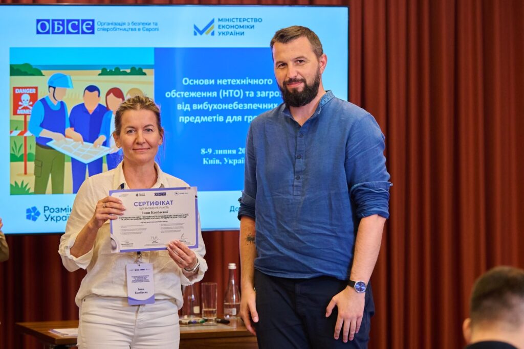 "Двоє людей на події в Києві, Інна Колбаєва тримає сертифікат. На екрані за ними — інформація про тренінг з нетехнічного обстеження та вибухових загроз для цивільних. Видно логотипи ОБСЄ та Мінекономіки України.