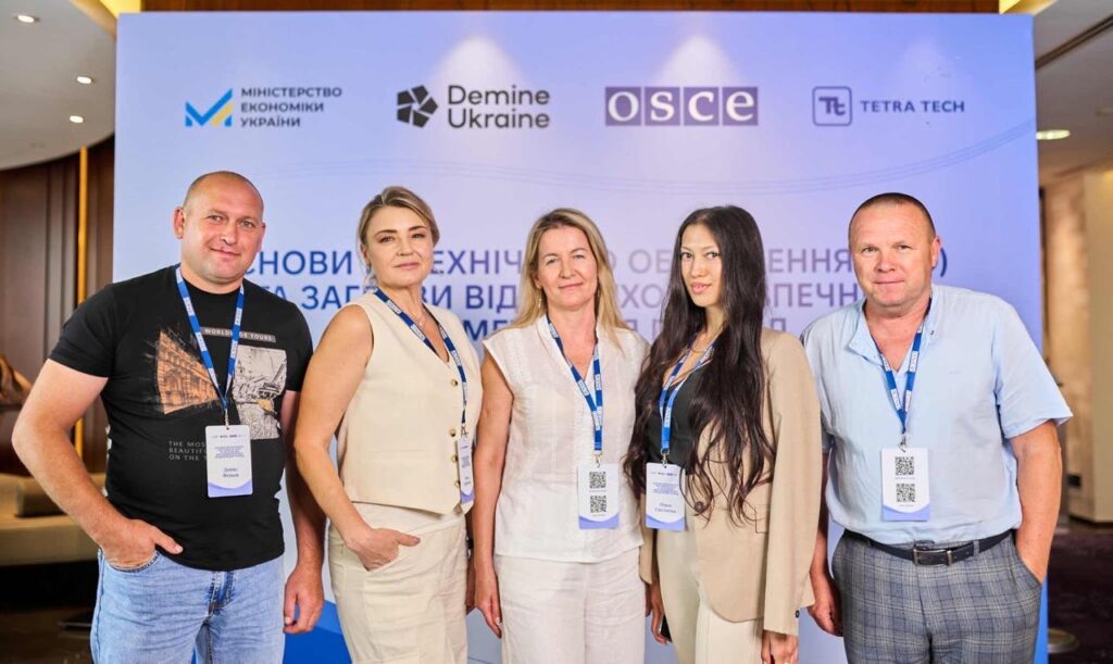 П’ятеро людей стоять перед банером з логотипами Міністерства економіки України, Demine Ukraine, ОБСЄ та Tetra Tech. 