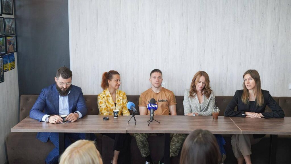 П’ятеро людей сидять за столом під час пресконференції або офіційної зустрічі. На столі — мікрофони (один із логотипом «UNN»), стаканчики з кавою, пластикові стакани з трубочками та мобільні телефони. Учасники вдягнені по-різному: діловий костюм, строката сорочка, футболка, піджак у клітинку та чорний піджак. Позаду, на лівій стіні, розміщені декілька картин у рамках. Атмосфера офіційна, але невимушена.