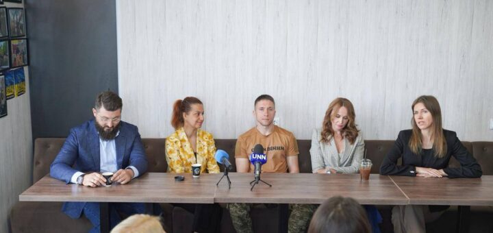 П’ятеро людей сидять за столом під час пресконференції або офіційної зустрічі. На столі — мікрофони (один із логотипом «UNN»), стаканчики з кавою, пластикові стакани з трубочками та мобільні телефони. Учасники вдягнені по-різному: діловий костюм, строката сорочка, футболка, піджак у клітинку та чорний піджак. Позаду, на лівій стіні, розміщені декілька картин у рамках. Атмосфера офіційна, але невимушена.