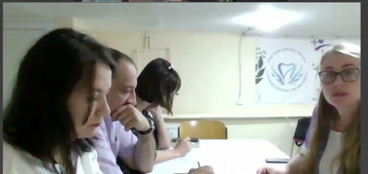 На зображенні — група людей за столом, які беруть участь у засіданні. На столі лежать документи та ручки. У верхній частині видно інтерфейс відеозв’язку, де зображені чотири учасники з підписами: «Олена Кириченко Управ...», «Наталя Устинова», «Ігор Савва» та «Музиківська СТГ». Трохи далі в кабінеті — банер із серцем і квітами та текстом. Унизу зображення — напис: «Кудрявцева Галина начальник відділу з питань ветеранської ХРД(ВА)».