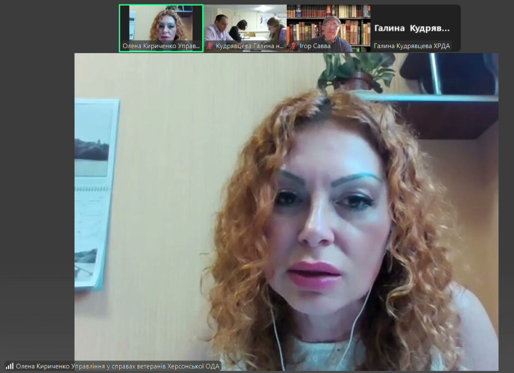 Скріншот відеоконференції, на якій видно п’ять учасників у окремих відеовікнах зверху. Всі учасники мають підписи під своїми вікнами, серед яких: «Олена Кириченко Управ...», «Кудрявцева Галина н...», «Ігор Савва», «Галина Кудряв...». У центральній частині — жінка з кучерявим волоссям у навушниках, яка сидить біля бежевої стіни. Зліва — настінний календар із зображенням, справа — полиця з вазоном та предметами. Внизу екрана — напис: «Олена Кириченко Управління у справах ветеранів Херсонської ОДА».