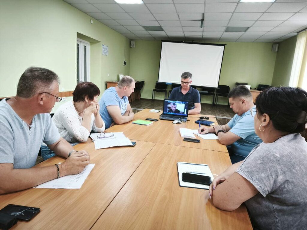 Засідання в кімнаті з блідо-жовтими стінами, де шестеро людей сидять за прямокутним конференц-столом. На передньому плані — ноутбук, відкритий на відеозв’язку, що поєднує присутніх із ще одним учасником дистанційно. На столі — зошити, ручки, смартфон, папери — усе свідчить про серйозну робочу атмосферу. Освітлення флуоресцентне, а на стіні розміщено великий екран для презентацій.