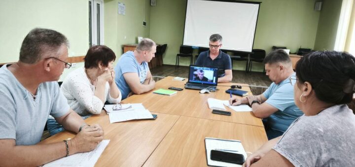 Засідання в кімнаті з блідо-жовтими стінами, де шестеро людей сидять за прямокутним конференц-столом. На передньому плані — ноутбук, відкритий на відеозв’язку, що поєднує присутніх із ще одним учасником дистанційно. На столі — зошити, ручки, смартфон, папери — усе свідчить про серйозну робочу атмосферу. Освітлення флуоресцентне, а на стіні розміщено великий екран для презентацій.