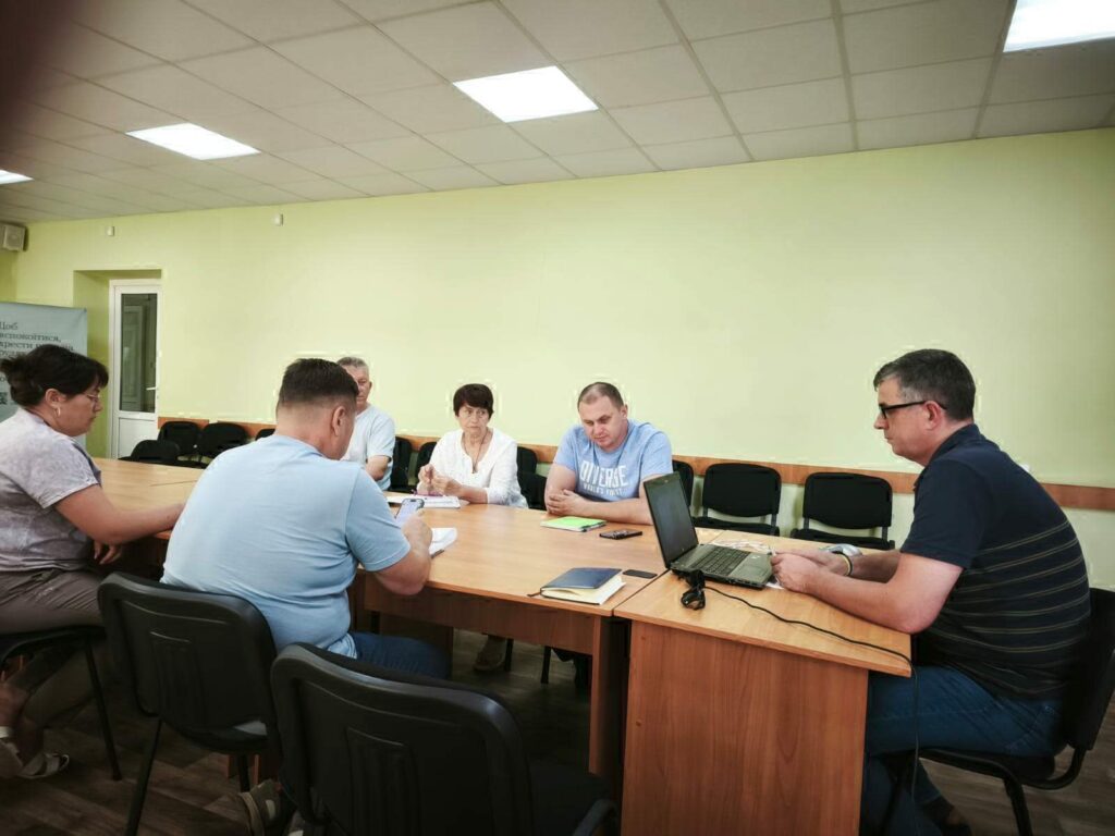 Шестеро людей за прямокутним столом у кімнаті з м’ятно-зеленими стінами. Перед Михайлом Линецьким стоїть ноутбук, на столі лежать зошити та папери, що створює атмосферу ділової зустрічі. У приміщенні видно двері, вікно та кілька додаткових стільців уздовж стіни