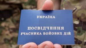 Рука тримає синю картку з текстом українською: «УКРАЇНА. ПОСВІДЧЕННЯ УЧАСНИКА БОЙОВИХ ДІЙ». На фоні — камуфляжна військова форма. Ця посвідка засвідчує статус людини, яка брала участь у бойових діях, і є офіційним документом в Україні