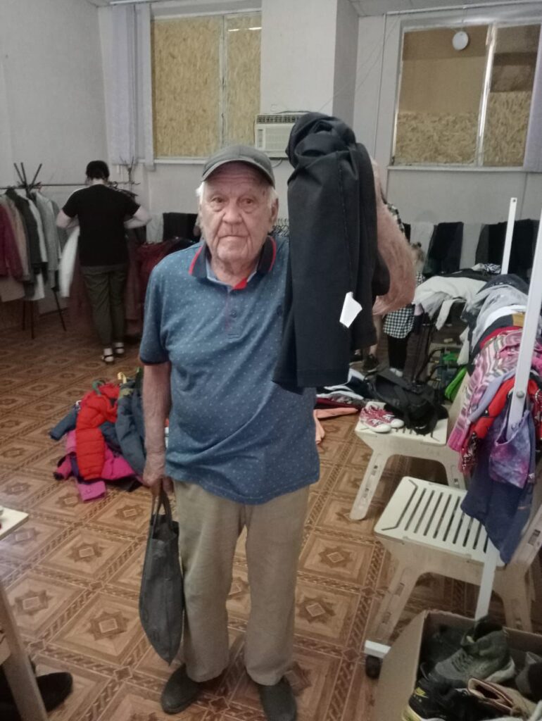 У кімнаті для видачі гуманітарної допомоги людина тримає в руках чорний одяг із білим цінником та сіру сумку. В приміщенні розміщено вішалки з різним одягом, частина речей лежить на стільцях і підлозі. Інша людина на задньому плані розглядає речі на вішалці. Підлога викладена плиткою з геометричним візерунком, на стінах — вікна з напівпрозорими фіранками та кондиціонер.