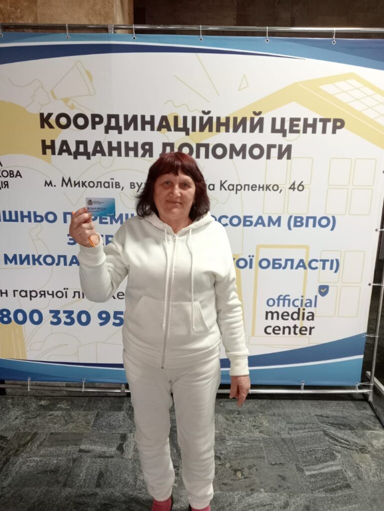 Жінка в білому спортивному костюмі та рожевому взутті стоїть перед великим банером. На банері — текст українською: «Координаційний центр надання допомоги, м. Миколаїв, вул. Карпенка, 46. Безкоштовно переміщеним особам (ВПО) Миколаївської області». У нижній частині банера написано «official media center», поруч вказано номер телефону: 800 330 95. Жінка тримає у лівій руці картку. Фото зроблено на вулиці або біля входу до будівлі.