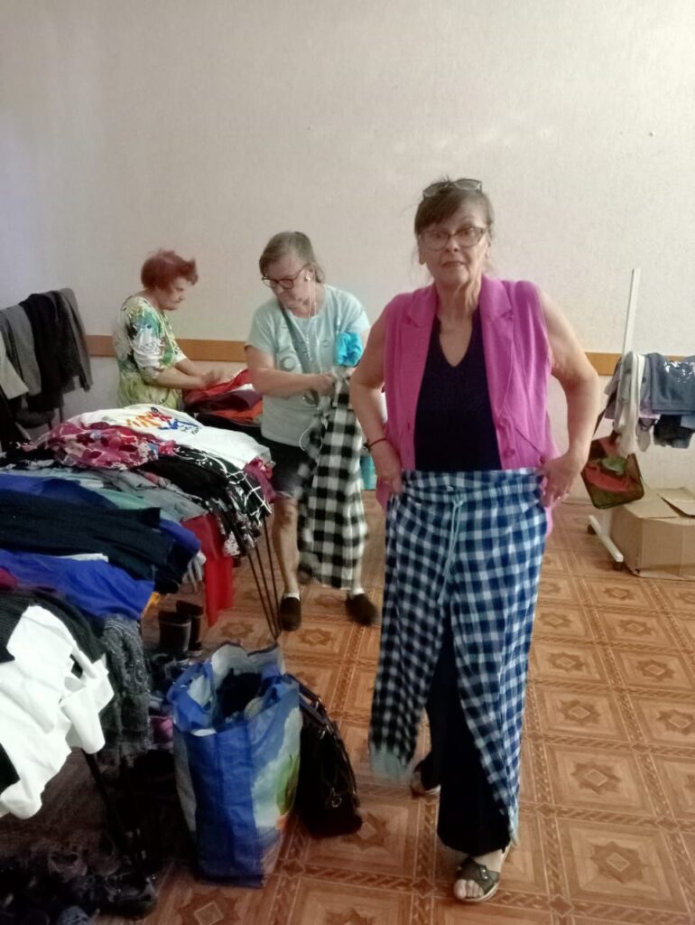 У кімнаті троє людей сортують одяг. На передньому плані — жінка в рожевій жилетці поверх темного одягу тримає в руках картату тканину синьо-білого кольору. На задньому плані інша людина тримає подібну тканину й розглядає речі. Ліворуч на столі — акуратно складений одяг, а на підлозі стоїть синя сумка з речами. Підлога вкрита плиткою з геометричним візерунком, стіни — однотонні. Обстановка нагадує пункт гуманітарної допомоги.