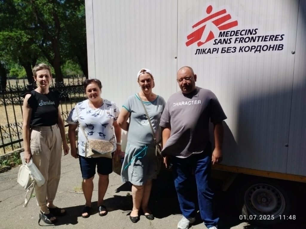 Чотири людини стоять перед мобільною конструкцією, на якій зображено логотип і назву організації Médecins Sans Frontières (Лікарі без кордонів) англійською та українською мовами. Логотип — стилізована фігура, що біжить. На конструкції також видно написи «Stardust» і «CHARACTER». Це схоже на мобільний медичний пункт або офіс підтримки. Усі стоять на асфальтованій території з огорожею та деревами на задньому плані. У нижній частині фото є позначка з датою та часом: 01.07.2025, 11:48.
