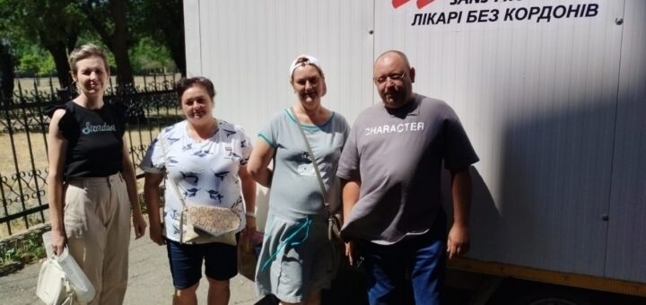 Чотири людини стоять перед мобільною конструкцією, на якій зображено логотип і назву організації Médecins Sans Frontières (Лікарі без кордонів) англійською та українською мовами. Логотип — стилізована фігура, що біжить. На конструкції також видно написи «Stardust» і «CHARACTER». Це схоже на мобільний медичний пункт або офіс підтримки. Усі стоять на асфальтованій території з огорожею та деревами на задньому плані. У нижній частині фото є позначка з датою та часом: 01.07.2025, 11:48.