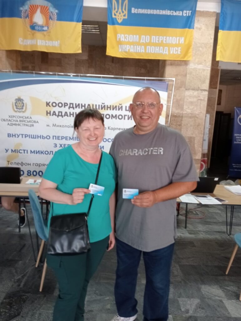 У приміщенні двоє людей стоять перед великим банером із текстом українською: «Координаційний центр надання допомоги внутрішньо переміщеним особам з Херсонської області у місті Миколаїв». Обоє тримають картки, ймовірно, пов’язані з отриманням допомоги або реєстрацією. На задньому плані видно два прапори: один із гаслом «Єдині разом!», другий — з написом «Разом до перемоги. Україна понад усе». Поряд стоїть стіл з ноутбуком і паперами. Атмосфера офіційна та підтримувальна.