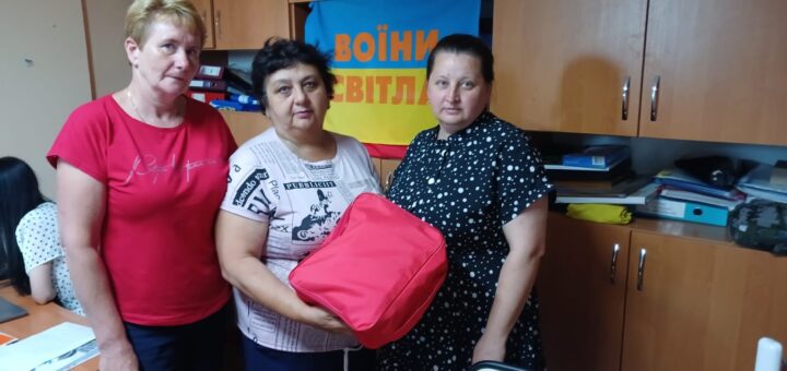 Троє людей стоять у кімнаті з дерев’яними шафами та полицями, заповненими папками, коробками й іншими офісними речами. Один із них тримає червону сумку - аптечку. Позаду — синьо-жовтий банер із написом українською: «ВОЇНИ СВІТЛА». Простір нагадує робоче приміщення або штаб підтримки. На столі ліворуч — документи та офісне приладдя