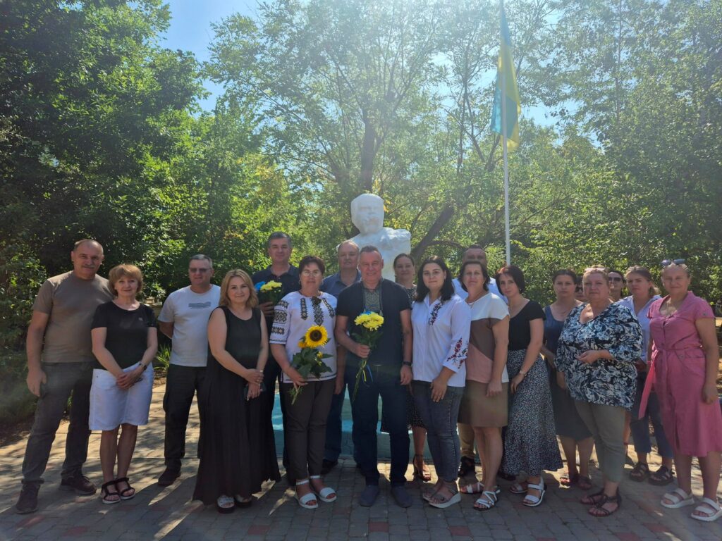 Група людей стоїть біля пам’ятника-бюста Т.Г. Шевченка на відкритому майданчику, поруч із флагштоком та прапором. Деякі учасники тримають квіти, зокрема соняхи. Довкола — дерева й зелень
