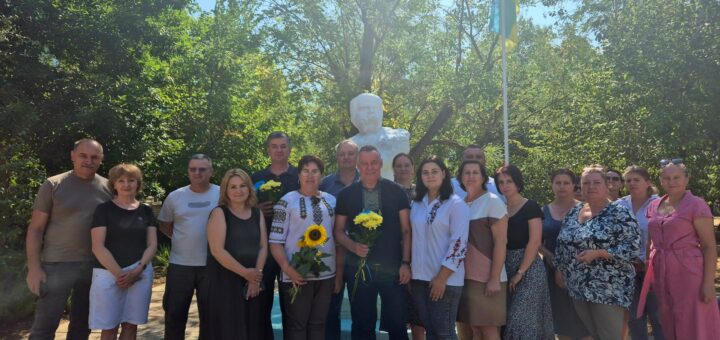 Група людей стоїть біля пам’ятника-бюста Т.Г. Шевченка на відкритому майданчику, поруч із флагштоком та прапором. Деякі учасники тримають квіти, зокрема соняхи. Довкола — дерева й зелень