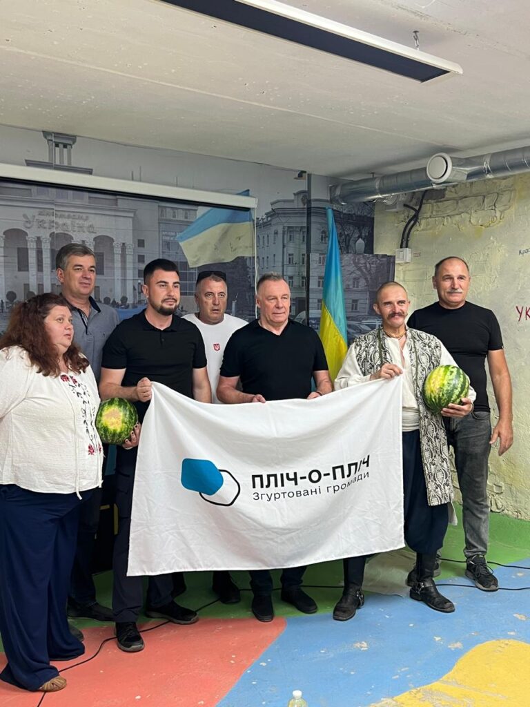 Група людей у приміщенні тримає білий банер із написом «ПЛІЧ-О-ПЛІЧ Згуртовані громади». Двоє учасників тримають кавуни. На фоні — український прапор і мурал із зображенням будинку та людей