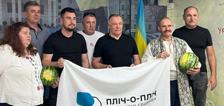 Група людей у приміщенні тримає білий банер із написом «ПЛІЧ-О-ПЛІЧ Згуртовані громади». Двоє учасників тримають кавуни. На фоні — український прапор і мурал із зображенням будинку та людей