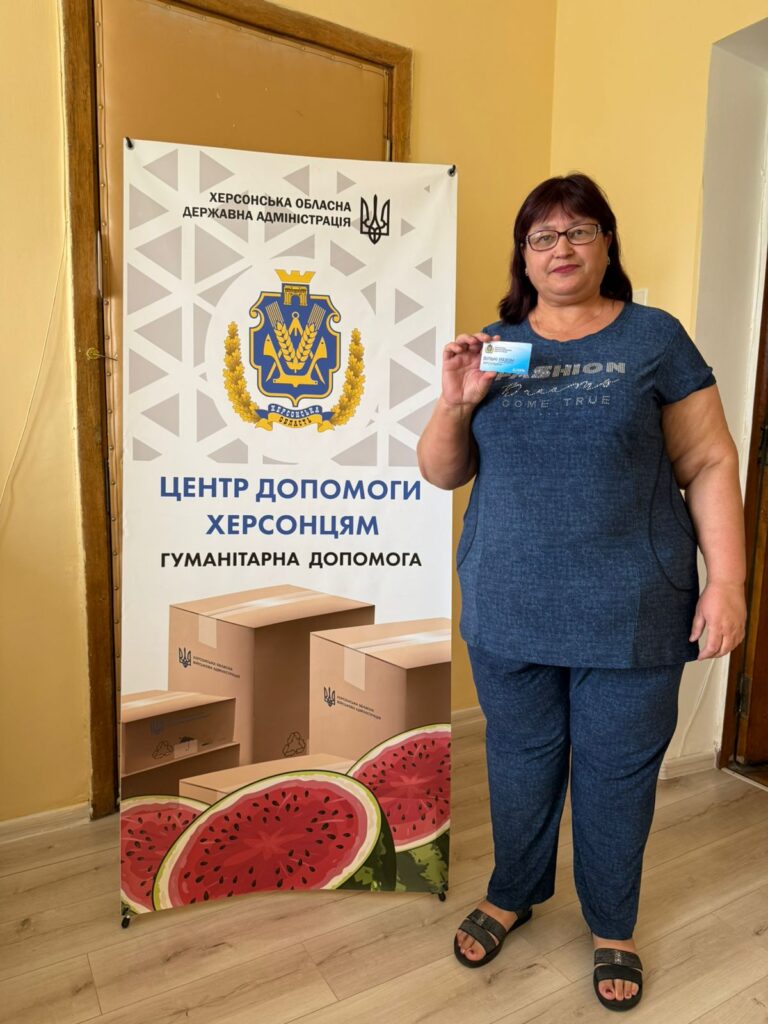 На зображенні — мешканка Ювілейного стоїть поряд із банером. На банері зображено герб Херсонської області та текст українською: «ХЕРСОНСЬКА ОБЛАСНА ДЕРЖАВНА АДМІНІСТРАЦІЯ», «ЦЕНТР ДОПОМОГИ ХЕРСОНЦЯМ», «ГУМАНІТАРНА ДОПОМОГА». У нижній частині банера — фото коробок та кавунів. Жінка тримає картку в правій руці.