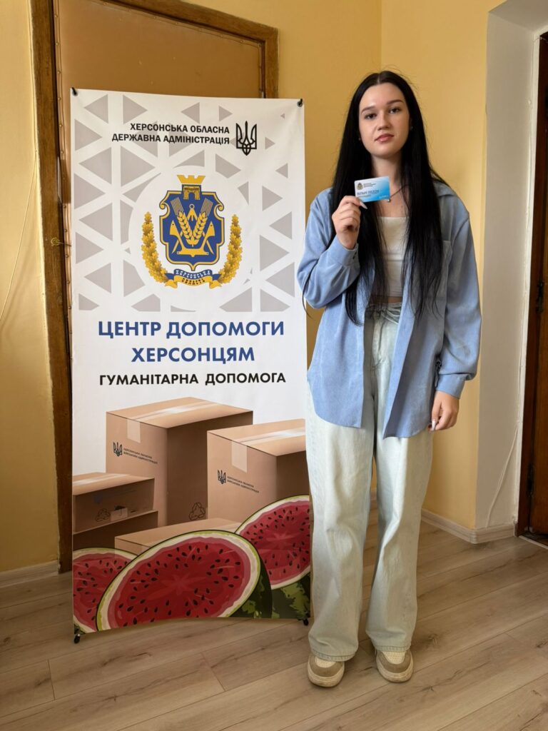 На зображенні — мешканка Ювілейного стоїть поряд із банером. На банері зображено герб Херсонської області та текст українською: «ХЕРСОНСЬКА ОБЛАСНА ДЕРЖАВНА АДМІНІСТРАЦІЯ», «ЦЕНТР ДОПОМОГИ ХЕРСОНЦЯМ», «ГУМАНІТАРНА ДОПОМОГА». У нижній частині банера — фото коробок та кавунів. Дівчина тримає картку в правій руці.