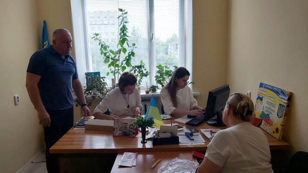 На зображенні — кімната з чотирма людьми, які працюють за столом. На підвіконні — кілька вазонів. На столі — комп’ютер, папери, канцелярське приладдя, маленький прапорець. Сергій Петроченко стоїть, троє співробітниць сидять за робочими столами. На стіні — плакат з текстом і зображеннями, пов’язаними з Україною.