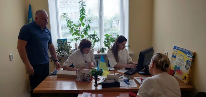 На зображенні — кімната з чотирма людьми, які працюють за столом. На підвіконні — кілька вазонів. На столі — комп’ютер, папери, канцелярське приладдя, маленький прапорець. Сергій Петроченко стоїть, троє співробітниць сидять за робочими столами. На стіні — плакат з текстом і зображеннями, пов’язаними з Україною.