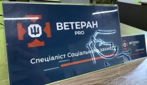 Як знайти фахівця з супроводу ветеранів На зображенні — стіл, на якому розміщено два таблички. Велика табличка містить текст українською: «ВЕТЕРАН PRO» та «Спеціаліст Соціального захисту», менша — «ВЕТЕРАН PRO» та «Світлана». На фоні — принтер, вазони та офісне обладнання.