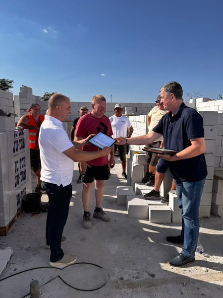 Група людей на будівельному майданчику серед білих бетонних блоків. Михайло Линецький вручає грамоту Коштурі Василю Омеляновичу. Позаду — кілька людей стоять або сидять на блоках, спостерігаючи. Ясний день із синім небом