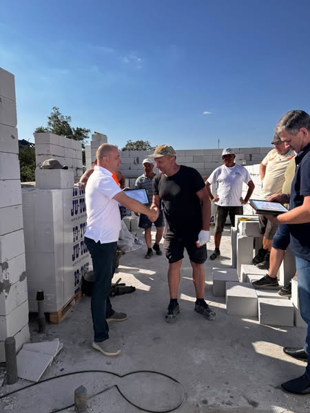 На будівельному майданчику двоє чоловіків тиснуть руки: один у білій сорочці, інший — у чорній футболці та шортах. Навколо — бетонні блоки, частково зведені стіни та будівельні матеріали. 