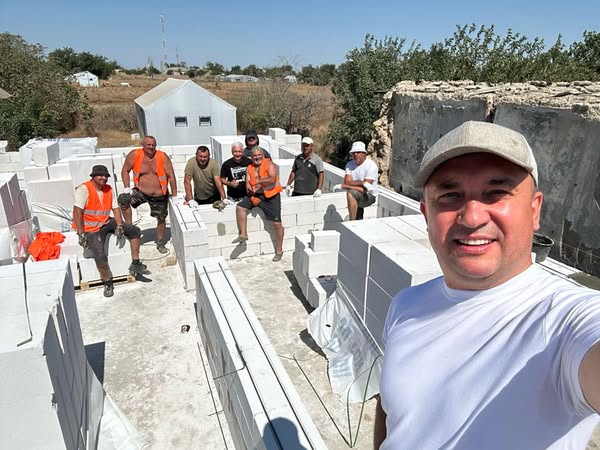 Вісім чоловіків на будівельному майданчику працюють над спорудженням із білих бетонних блоків. Деякі вдягнені в помаранчеві жилети, інші — в повсякденний одяг. Вони стоять або сидять на блоках, ніби позують для фото. На тлі — дерева та невелика біла будівля