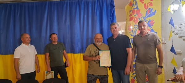 П’ятеро людей стоять перед синьо-жовтим тлом, схожим на прапор України. У центрі — чоловік тримає в руках рамку з сертифікатом або документом. Праворуч — стіна з квітковим орнаментом і трикутними прапорцями. Атмосфера урочиста, ймовірно, нагородження або офіційна подія.