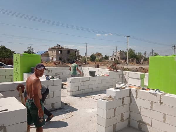 Будівельники зводять стіни з білих бетонних блоків на відкритому майданчику. Позаду — старі будівлі та електролінії. Сцена нагадує процес відновлення або будівництва в сільській чи приміській місцевості