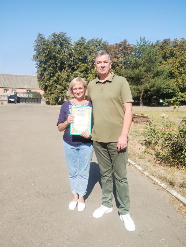 Михайло Линецький стоїть на вулиці поруч із жінкою, яка тримає Подяку. На задньому плані — дерева та будівля. Обоє вдягнені неформально, атмосфера невимушена.